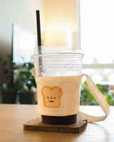 The canvas drink holder with bread patch design holding a plastic cup of coffee displayed on a wooden table from a different perspective
