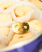 Shiny gold-colored hard enamel pin with metal butterfly clasp in the form of a pretzel cookie displayed in a tray of cookies