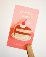 Chocolate cake slice with cherries and cream acrylic magnet displayed with a pink backing card on a white background.